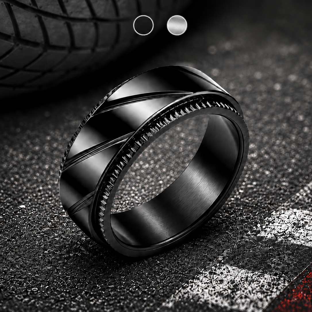 A black metallic spinning ring from Anxiety Rings Australia with a textured, ribbed outer band and a smooth brushed inner band, resting on a dark asphalt surface with a blurred tire in the background.