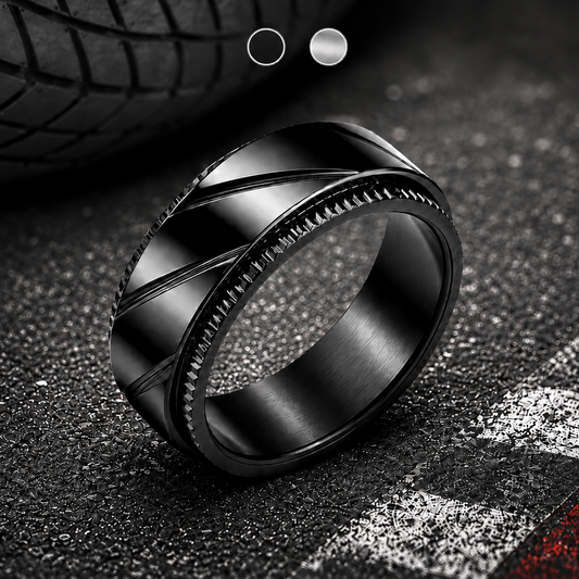 A black metallic spinning ring from Anxiety Rings Australia with a textured, ribbed outer band and a smooth brushed inner band, resting on a dark asphalt surface with a blurred tire in the background.