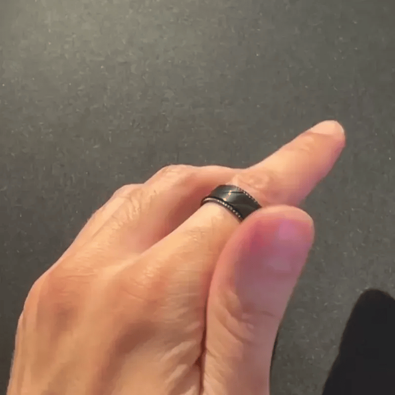 A close-up of a person's hand wearing a black band spinning ring from Anxiety Rings Australia with a textured center and small metallic edge details, set against a dark gray background.