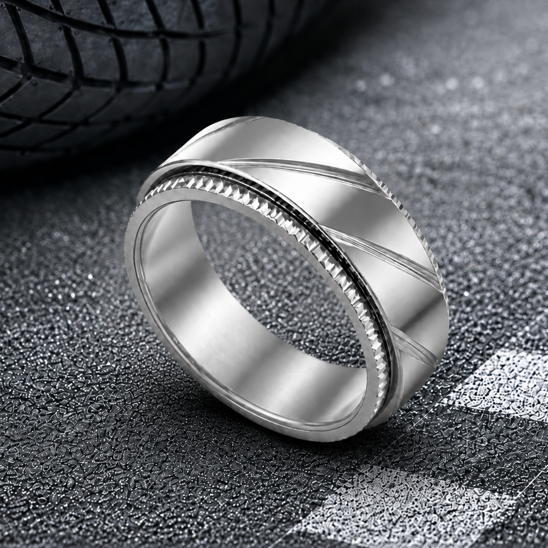 A silver-toned metal band spinning ring from Anxiety Rings Australia with a textured diagonal pattern and a beaded channel detail, displayed on a dark asphalt surface with a tire in the background.