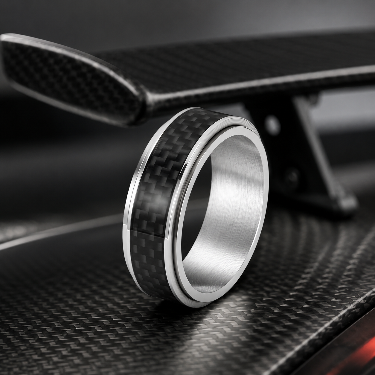 A carbon fibre spinning ring from Anxiety Rings displayed on a piece of carbon fibre with a carbon fibre car wing in the background