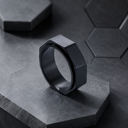A dark metallic hexagonal spinning ring from Anxiety Rings Australia standing upright on a textured gray stone pedestal, with a matching dark gray hexagonal tile pattern in the background.