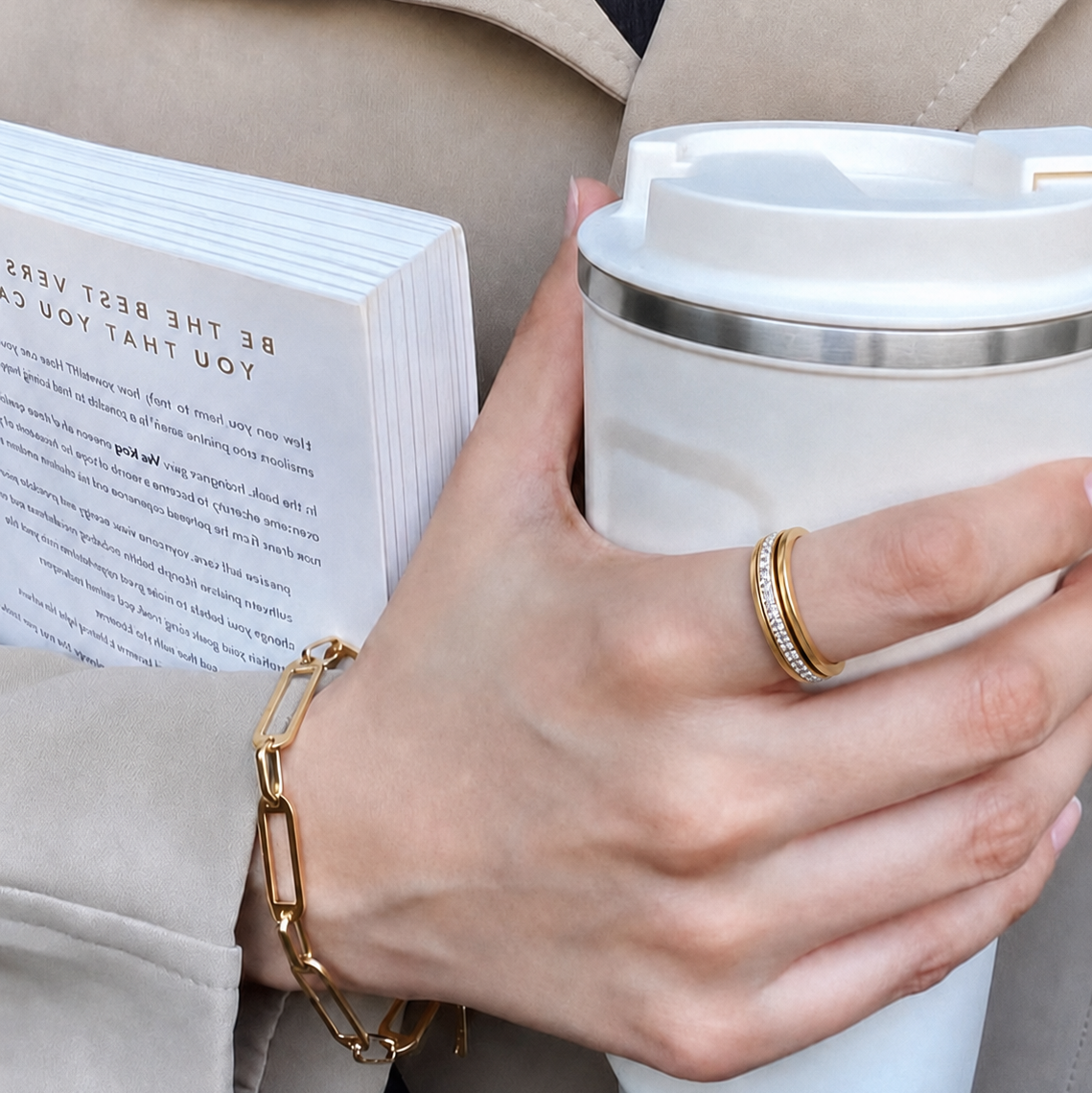 A close-up lifestyle shot of a person holding a white travel mug and a book, wearing a gold chain bracelet and a gold lumiere spinning ring from anxiety rings australia, on index finger.