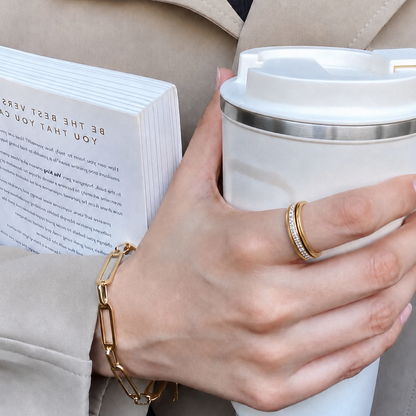 A close-up lifestyle shot of a person holding a white travel mug and a book, wearing a gold chain bracelet and a gold lumiere spinning ring from anxiety rings australia, on index finger.