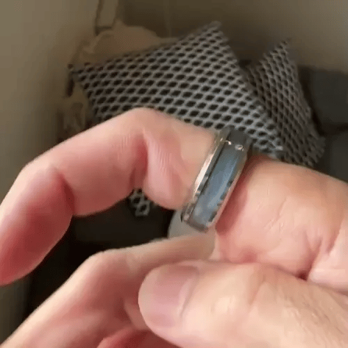 A close-up gif of a person's hand wearing a silver-toned spinning ring from Anxiety RIngs Australia with a light blue inlay, set against a blurred indoor background with patterned pillows.