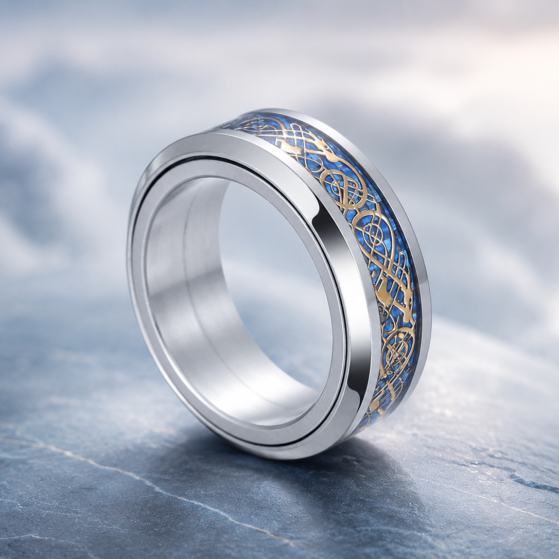 A polished silver-toned spinning ring from Anxiety RIngs Australia with an intricate blue and gold Celtic knot pattern inlay, displayed on a textured gray stone surface with a soft, blurred cloudy background.