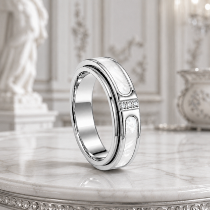 A polished silver spinning ring from Anxiety Rings Australia with white inlay panels and a row of small clear stones, displayed on a marble surface in an ornate interior setting.