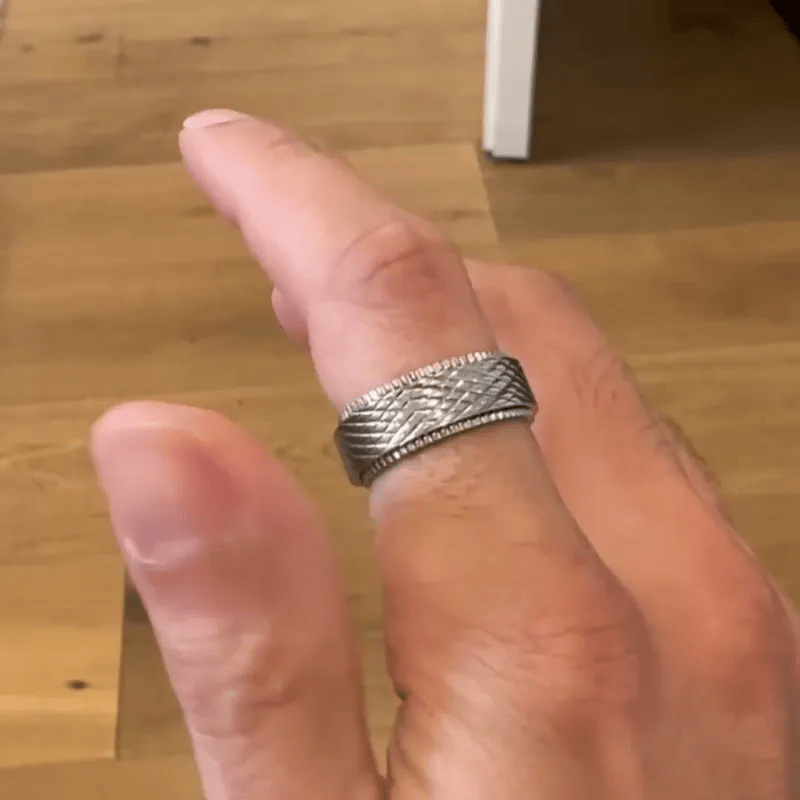 A silver textured band A silver-toned textured spinning ring from Anxiety Rings Australia with a diamond-patterned band and a beaded inner edge, displayed on a dark textured scale-like surface. worn on a person's finger, shown against a light wood floor background.