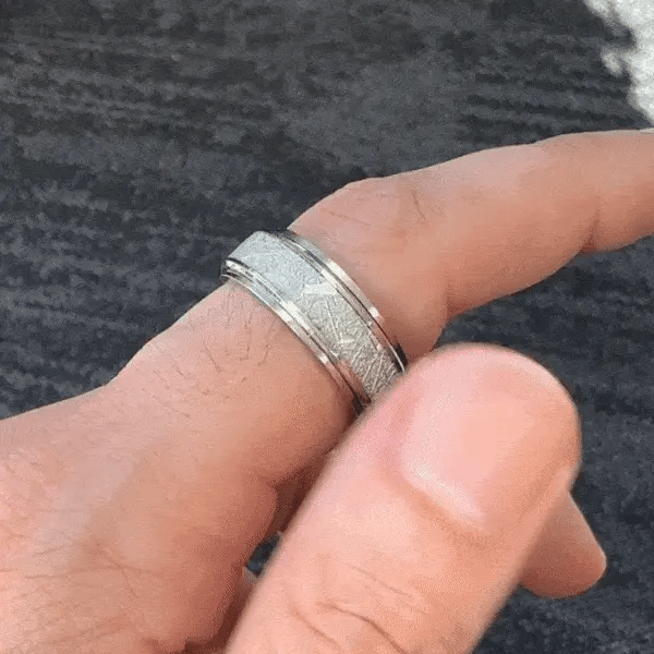 A close-up gif of a person's hand wearing a silver-toned spinning ring from Anxiety RIngs Australia with a light blue inlay, set against a blurred indoor background with patterned pillows.